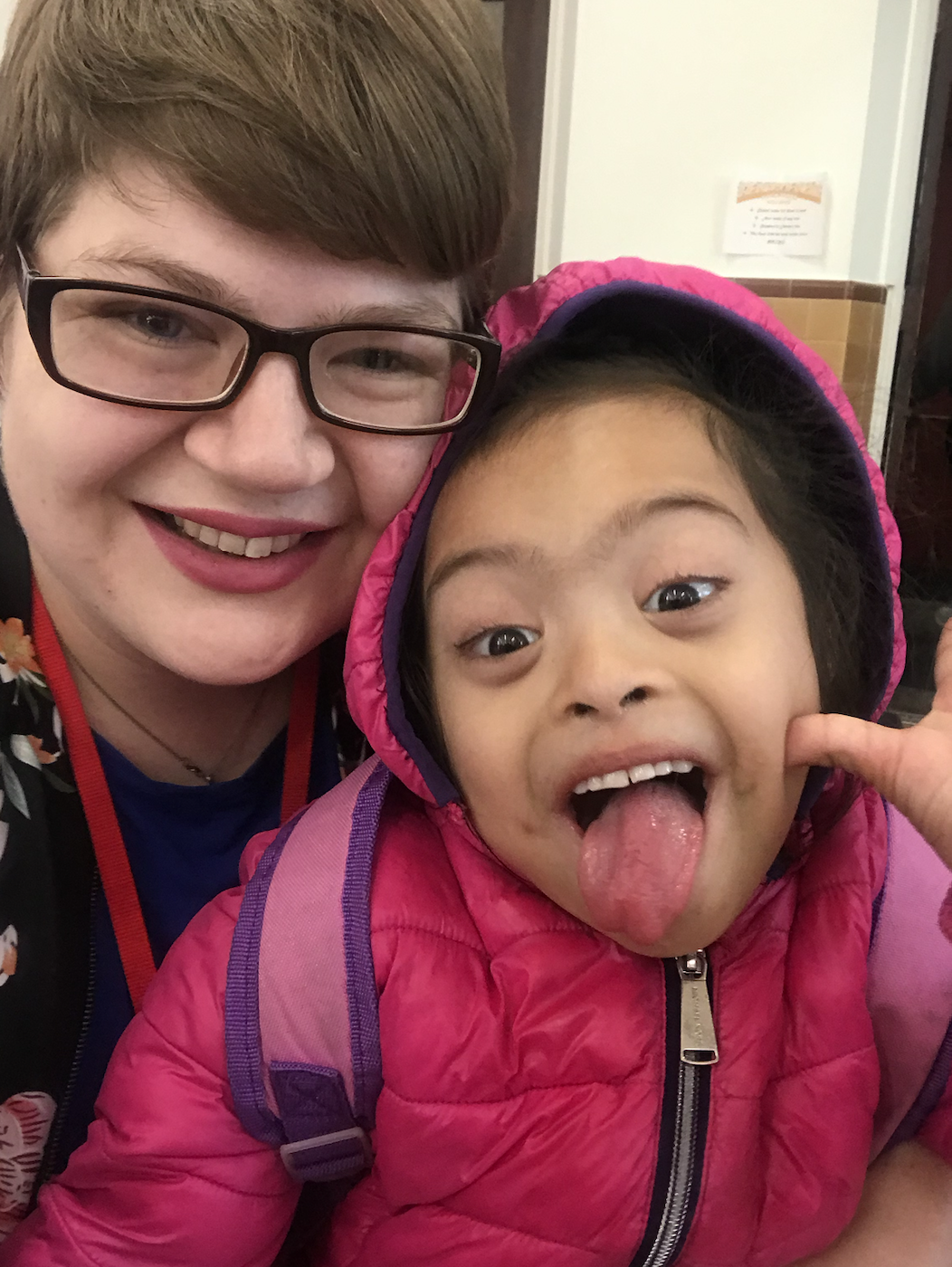 a selfie image of a smiling, white, non-binary adult with glasses and a latina child in a bright pink coat with her tongue sticking out in a playful gesture